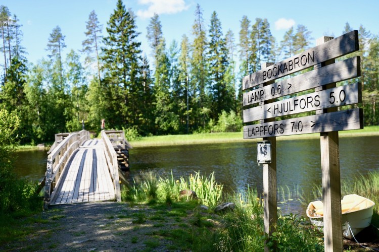 Pitkä silta joen yli. Joen varrella opastetaulu, jossa mainitaanétäisyydet eri kohteisiin. 