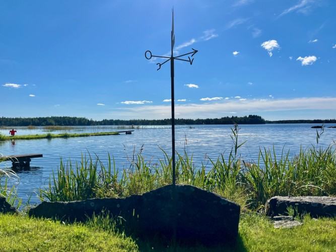 En gräsmatta vid en sjö. I sjön finns en brygga. På gräset är en stor sten och en väderstreckvisare.