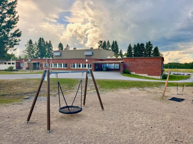 Edsevön koulupiha. Etualalla hämähäkkikeinu, taustalla koulurakennus. Dramaattiset pilvet koulun yllä.