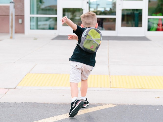 En pojke med skolväska på ryggen framför en skolbyggnad.