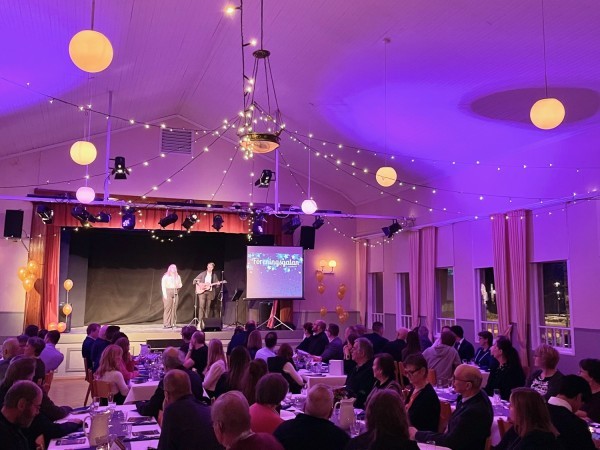 Människor sitter till bords i en festligt inredd lokal. På scenen står två personer som sjunger och spelar gitarr.