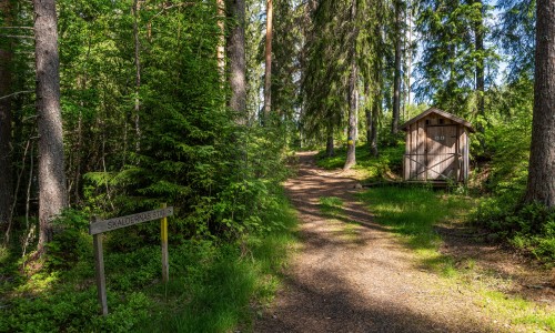 Leveä polku metsässä ja oikealla ulkohuussi. Kyltissä lukee: Runoilijoiden polku.