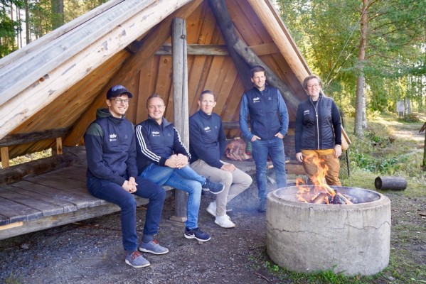 Tre män sitter i ett vindskydd, en man och en kvinna står bredvid. Framför dem brinner elden i en rund cementring.