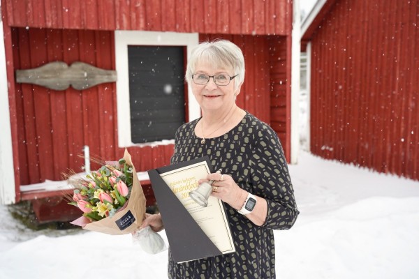 Gunhild Berger seisoo omalla pihalla, talvimaisemassa. Hänellä on kukkakimppu, kulttuuripalkinto Pedersöre-kello ja diplomi kädessään.