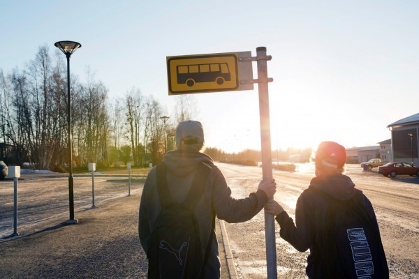 Motsolsbild. Två unga personer står och håller i en stolpe på en busshållsplats.