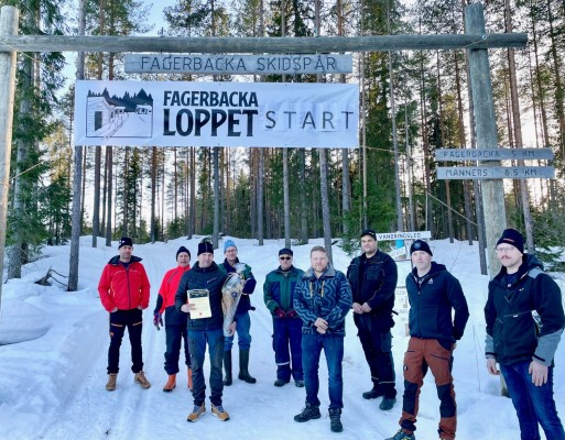 Yhdeksän ihmistä seisoo hiihtoladun lähtopisteessä, Fagerbackalatukyltin ja hiihtokilpailu Fagerbackaloppetin mainostavan kyltin alla. 