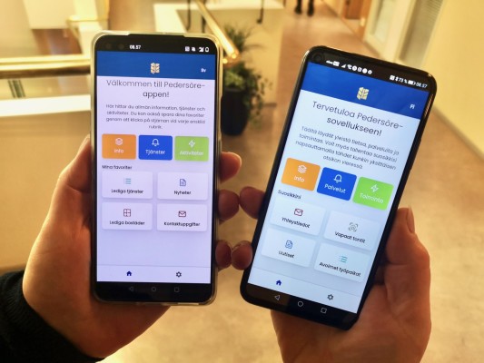 Kaksi mobiilipuhelinta, joissa Pedersöre-sovellus näkyy näytöllä. Sekä ruotsin- että suomenkielinen sovellusversio.