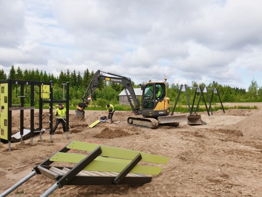Lekparken byggs med grävmaskin på en sandig plan. Det ligger halvfärdiga lekredskap på marken.