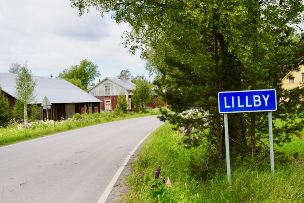 Maantie kulkee omakotialueen läpi. Kuvan oikealla puolella on kyltti tekstillä LILLBY. On kesä.