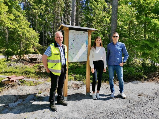 Tre personer står bredvid en skylt, två karlar och en dam i mitten. Skylten visar en karta över Sandsund vandringsled.