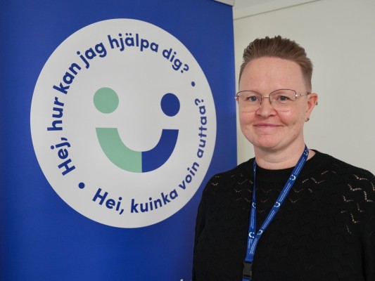 Camilla Passell står bredvid en skylt med Österbottens välfärdsområdes logo. På logon står det Hej, hur kan jag hjälpa dig? Hei, kuinka voin auttaa?