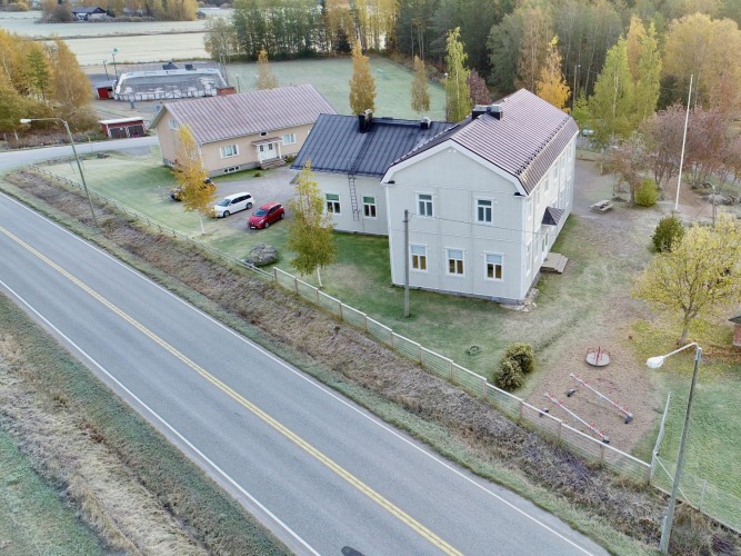 Droonikuva entisesta koulurakennuksesta, vuokratalosta, koulun pihasta ja tontin ohi kulkevasta maantieltä.