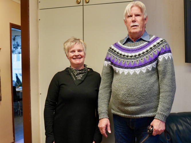 Nainen ja mies seisovat vierekkäin ja hymyilevät kameraan. He ovat sisätiloissa. Taustalla näkyy toinen huone. Mies on huomattavasti naista pidempi.