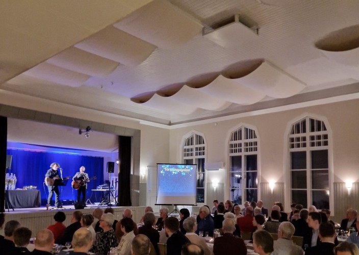 Kuva aiemmasta seuragaalasta. Kaksi esiintyjää ison salin lavalla. Ihmiset istuvat pitkän pöydän ääressä.