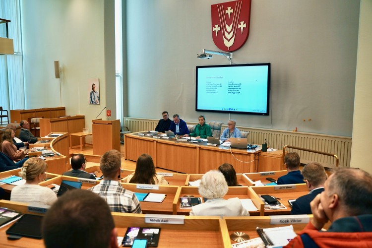 Kunnanvaltuuston istuntosali. Puhujakorokkeella istuu valtuuston puheenjohtajisto. Kokous on käynnissä.