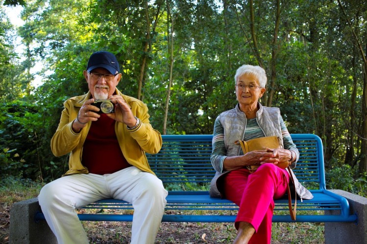 Två seniorer, man och kvinna, sitter på en bänk. Mannen har en kamera.