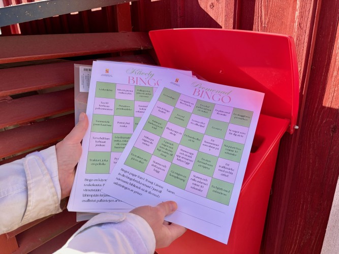 En person håller i bingobrickorna, en svenskspråkig och en finskspråkig, vid en motionslåda.