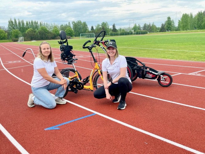 Rörelsecoacherna Ida NIemi och Julia Byggmästar står på knä på löparbanorna i Bennäs. I bakgrunden vagn och cykel anpassad för personer med funktionsnedsättning.