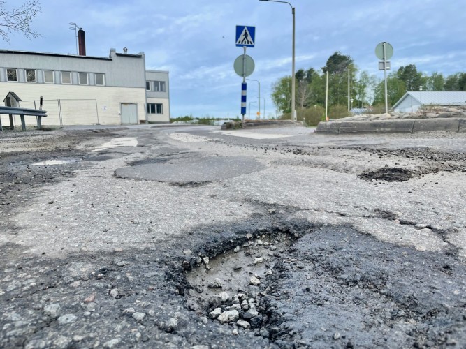 Gropar och ojämn väg. En byggnad syns i bakgrunden.