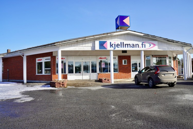 Fastigheten som mopedverkstaden placeras i. Huvuddörr och fastighetens framsida. Ovanför huvuddörren står det "Kjellman.fi". En parkerad bil till höger. 