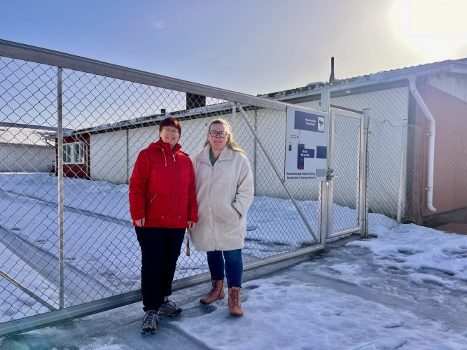 Två kvinnor står framför en stängd metallgrind. Innanför inhägnaden finns en stor fastighet i tegel.