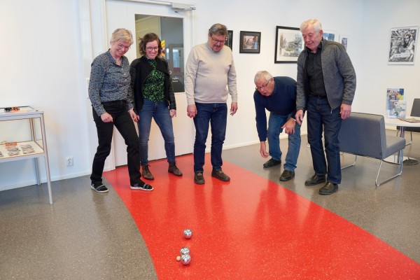 Fem personer står i Galleri Bruno. Fyra ser på när en av männen i bild kastar ett bouleklot mot den lilla bollen som används i boulespel. Damen längst till vänster knyter näven i luften som i triumf.