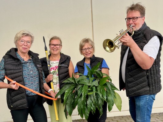 Medborgarinstitutets personer står på rad och håller i olika saker: Rektorn håller i en hulahularing, textilläraren har en innebandyklubba, kurssekreteraren har en grönväxt och musikläraren en trumpet.