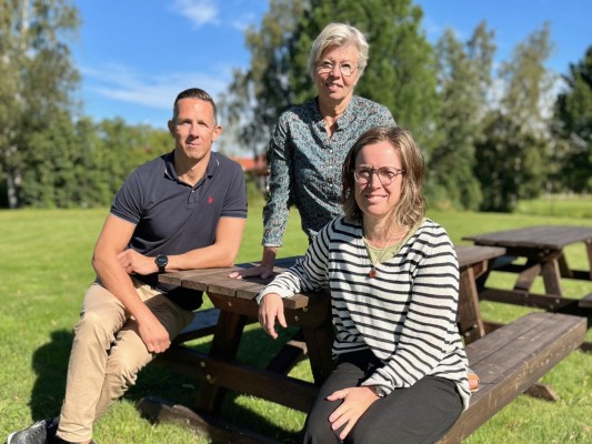 Liikuntasihteeri Pontus Backlund, kansalaisopiston rehtori Pia Boström ja kulttuuri- ja hyvinvointipäällikkö Ulrika Stenmark istuvat puupenkillä Kulttuuritalo Axin vieressä. Aurinko paistaa.