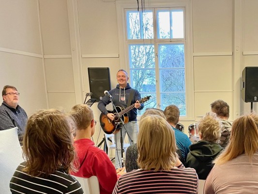 Körövning, där körledaren Dennis står framför kören. Han håller i en gitarr. Mikael sitter vid pianot och spelar.