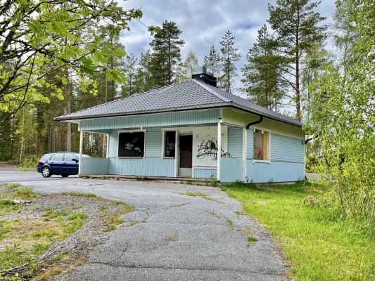 Huset som ska rivas omgiven ev vårgrönska. Framför kiosken står en parkerad bil.