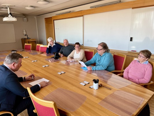 Kunnanhallituksen huoneessa istuu kuusi henkilöä. Kunnanjohtaja istuu yksin vasemmalla, Lappforsin edustajat istuvat pöydän oikealla puolella. Kyläneuvoston puheenjohtaja lukee kirjelmän.
