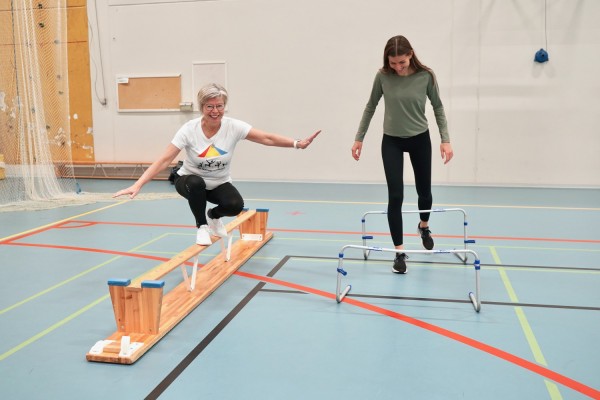 Pia Boström står på en upp-och ner-vänd bänk medan Jennifer Storrank går över små hinder i sportishallen.