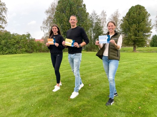 Motionsrådgivaren Jennifer, idrottssekreteraren Pontus och kanslisten Camilla står på gräsmatta och håller varsitt kort i händerna.