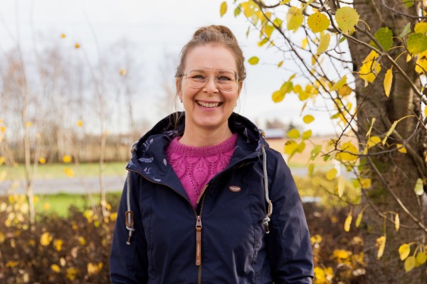 Anna-Karin Pensar fotograferad utomhus i höstlik miljö. Hon ler stort mot kameran.