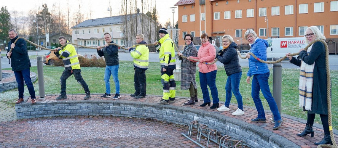 Kymmenen ihmistä seisoo rivissä ja pitää yhtä köyttä hartioillaan. He "työskentelevät yhdessä".