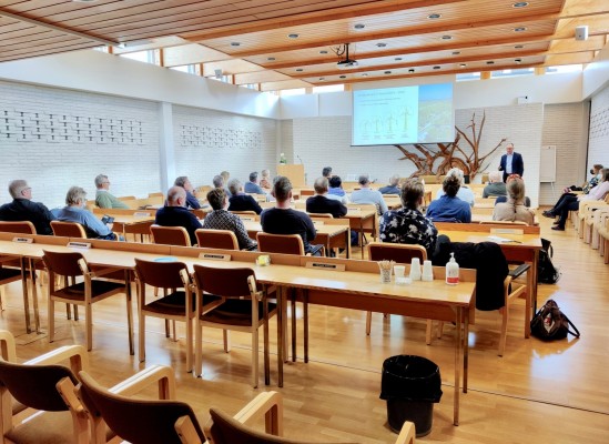 En stor sal, en man står längst fram och håller en presentation. Besökarna från Pedersöre sitter vid långa bord och lyssnar.