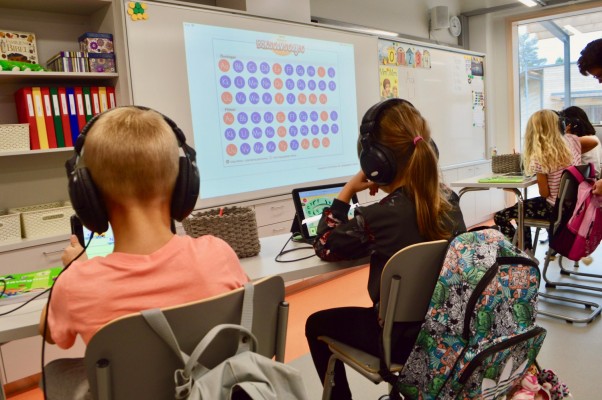 Två elever sitter i ett klassrum. Vi ser dem bakifrån. Framme i rummet finns en tavla.