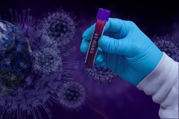 En hand håller i ett provrör med blod. I bakgrunden coronavirusbollar.
