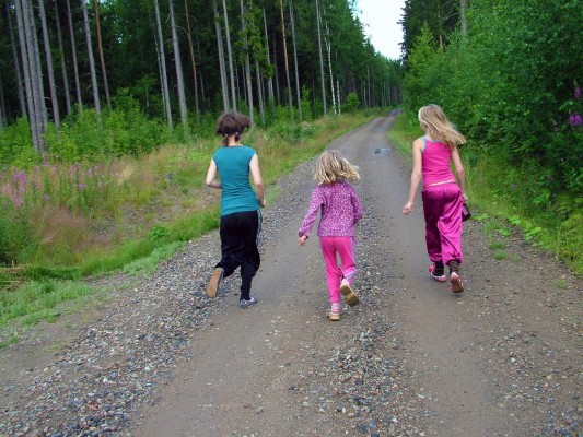 Kolme lasta juoksee hiekkatiellä metsässä.