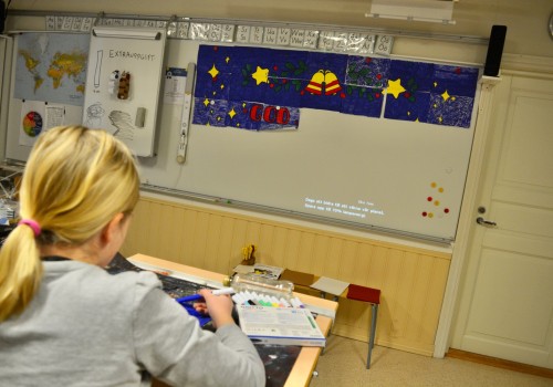 Ryggen av en elev som sitter i ett klassrum och klipper och klistrar. Längst fram i klassen finns mörkblåa papper på tavlan.