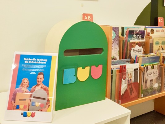 BUU-klubbenin postilaatikko, jolla on kolme sydäntä etusivulla. Postilaatikon viereen on kyltti, jossa on tietoa postilaatikon toiminnasta. Kuvan oikeassa reunassa näkyy kirjoja.