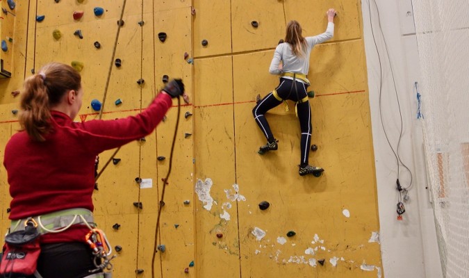 En person klättrar på klätterväggen i Sportis. En person står på golvet och säkrar, med linor.