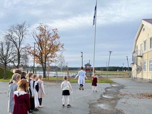 Elever iklädda gammaldags kläder, hoppar långrep på skolgården. Finlands flagga är hissad i flaggstången.
