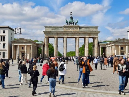 En bild från ett tog i Berlin. Det går många personer på torget och i bakgrunden syns ett monument, som består av pelare och ett tak.
