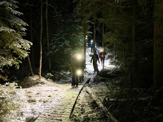 Det är mörkt och snö på marken i en skog. Man ser några personer som går på en stig. De har pannlampor som lyser upp marken.