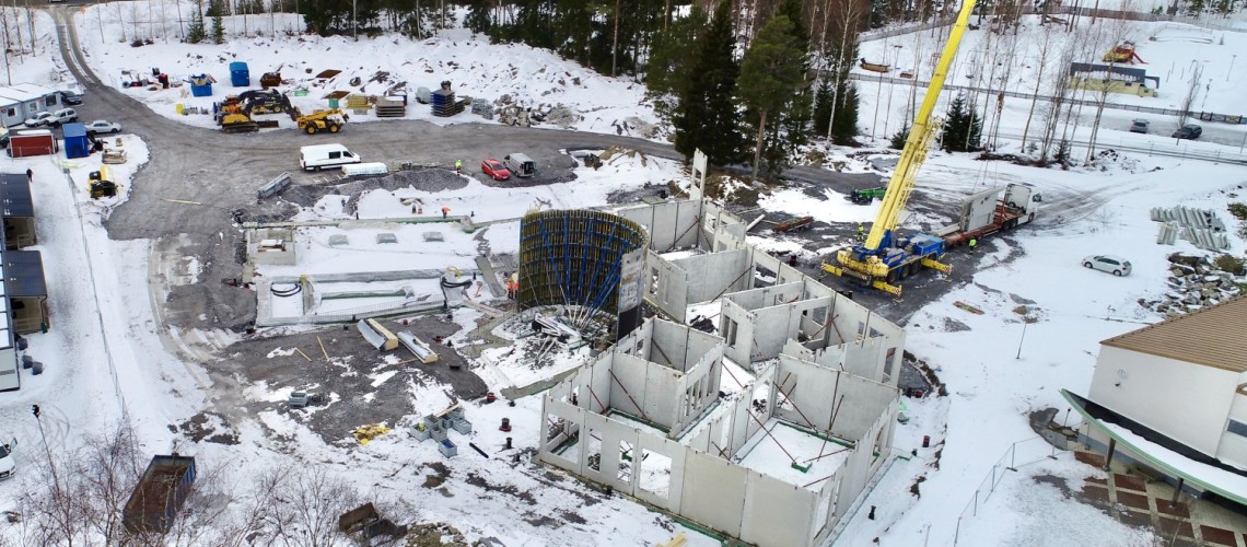 En flygbild över området där skolan byggs. En lyftkran, elementväggar som lyfts på plats, arbetsmaskiner och baracker. Det är snö på marken.