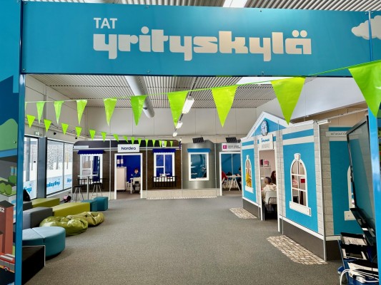 Bild från Företagarbyn. En inomhusmiljö med kontor, trekantiga flaggor hänger över mittgången, en skylt med texten Yrityskylä (sv Företagarbyn).