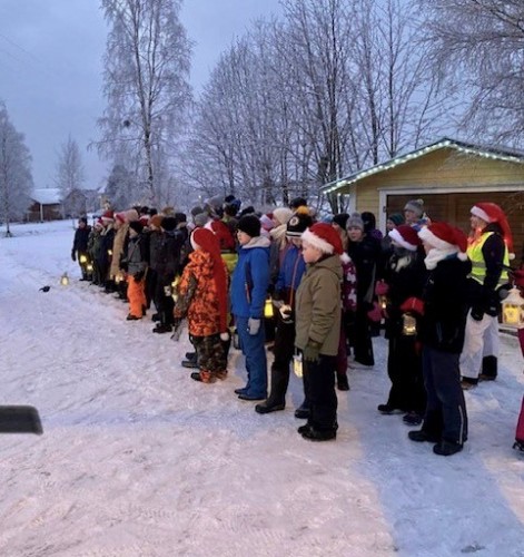Barn på en lång rad i vinterlandskap. De är klädda i tomteluvor och har lyktor i händerna.