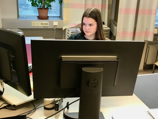 Teini-ikäinen pitkätukkainen tyttö istuu pöydän ääressä ja työskentelee tietokoneella.