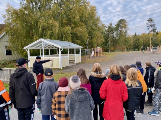 En grupp med elever lyssnar medan läraren berättar och pekar in mot skolgården.